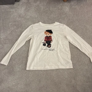 Vit långärmad tröja Polo Ralph Lauren - Vit långärmad tröja från Polo Ralph Lauren med ikonisk Polo Bear-tryck på bröstet. Björnen har en rutig röd jacka och svart basker, vilket ger en cool och lekfull vibe. Tröjan är gjord i mjuk bomull och har en klassisk passform som funkar till många stilar.