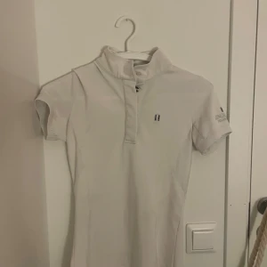 Vit pikétröja Kingsland Equestrian - Snygg vit pikétröja från Kingsland Equestrian med korta ärmar och ståkrage. Tröjan har diskret logga på bröstet och broderad text på ärmen. Materialet är mjukt och andas, perfekt för ridning och sommar. Passformen är normal och modellen är klassisk.