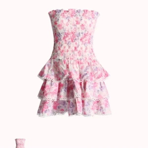 Blommig volangklänning från design by Amor - Supersöt kort klänning med blommigt mönster i rosa, lila och vitt. Axelbandslös och med smock upptill, samt två lager volanger med spetskant. Perfekt för sommarens alla tillfällen! Materialet känns lätt och luftigt.