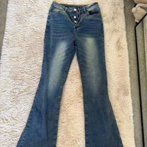 Blå bootcut jeans med knappar - Snygga blå bootcut jeans med hög midja och knappar framtill. Perfekta för en avslappnad stil med en touch av retro. De har en klassisk femficksdesign och en lätt tvättad look.