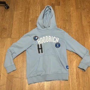 Ljusblå hoodie från Hoodrich - Säljer en stilren ljusblå hoodie från Hoodrich med tryck och broderade detaljer på framsidan. Hoodien har en stor ficka framtill och en bekväm huva. Perfekt för en avslappnad stil.