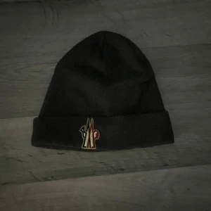 Svart mössa från Moncler - Snygg svart mössa från Moncler med broderad logotyp framtill. Perfekt för att hålla värmen under kyliga dagar. Klassisk design med uppvikt kant.
