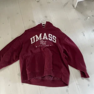 Vinröd college tröja - Säljer en vinröd hoodie med stort UMass-tryck på framsidan. Tröjan har en klassisk känguruficka och en bekväm huva. 