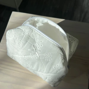 Vit quiltad necessär - Säljer en söt vit quiltad necessär med broderade blommor och en liten rosett på framsidan. Perfekt för att förvara smink eller andra småsaker. Den har en praktisk dragkedja upptill för enkel åtkomst.