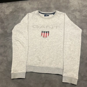 Grå sweatshirt från GANT - Säljer en stilren grå sweatshirt från GANT med ett broderat emblem på bröstet. Tröjan har rund hals och långa ärmar, perfekt för en avslappnad look. Passar bra till både jeans och chinos.
