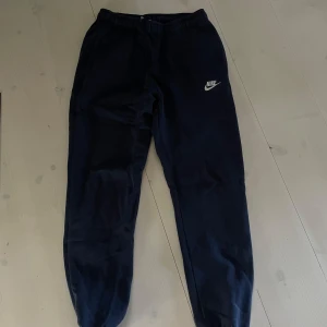Marin blåa mjukisbyxor från Nike - Säljer ett par blåa mjukisbyxor från Nike med elastisk midja och logga på benet. Perfekta för en avslappnad stil eller träning. Byxorna är tillverkade i mjukt material för extra komfort.