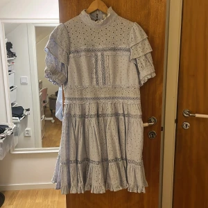 Ljusblå klänning med spetsdetaljer - Charmig ljusblå klänning med spetsdetaljer och volanger. Klänningen har korta ärmar och en hög hals. Perfekt för en somrig look med sin luftiga design och fina detaljer.