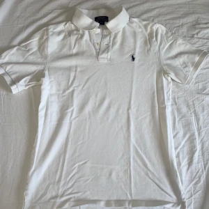 Vit pikétröja från Ralph Lauren - Säljer en klassisk vit pikétröja från Ralph Lauren med korta ärmar och den ikoniska loggan broderad på bröstet. Storlek L i barnstorlek. Sitter som en S