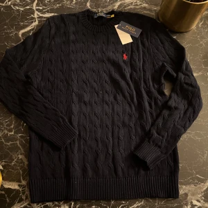 Marinblå stickad tröja från Polo Ralph Lauren - Säljer en stilren marinblå stickad tröja från Polo Ralph Lauren med kabelstickat mönster. Tröjan har en broderad röd logotyp på bröstet och ribbstickade muddar vid ärmslut och nederkant. Perfekt för en klassisk look.