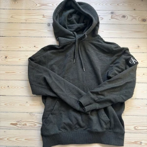 MörkGrön hoodie från Calvin Klein - Säljer en stilren mörkgrön hoodie från Calvin Klein. Den har en klassisk design med en justerbar huva och ribbade muddar. Perfekt för en avslappnad look. CK-logga på ärmen ger en extra touch av stil. Skick 8/10