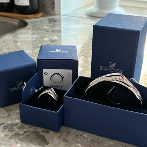 Elegant set med ring och armband i silverfärg  - Snyggt set med en ring och ett armband i silver. Båda smyckena har en modern design med flera tunna band som ger en stilren look. Perfekt för att ge en touch av elegans till din outfit. Från Swarovski, köpta på deras hemsida. Använda fåtal gånger, ringen är lite för stor för mig. Storlek M i armbandet och storlek 60 i ringen 
