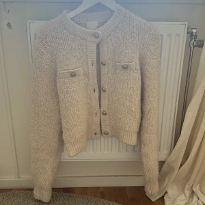 Beige fluffig kofta från H&M - Mysig beige kofta från H&M med dekorativa knappar och två fickor framtill. Koftan har en mjuk och fluffig textur, perfekt för kyliga dagar. Den har långa ärmar och en rund halsringning. Perfekt för att hålla sig varm och stilren.