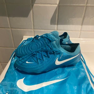Nike Phantom Elite AG - Nike phantom elite. blå fotbollsskor från Nike med vit logga och snörning. Skorna har en modern design med dobbar för optimalt grepp på planen. Perfekta för fotbollsspelare som vill ha stil och funktionalitet. 