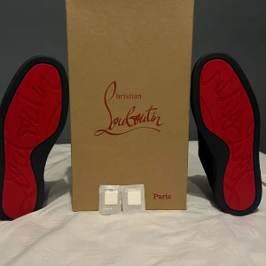 Svarta sneakers från Christian Louboutin - Snygga svarta sneakers från Christian Louboutin med den ikoniska röda sulan. Skorna har en stilren design med snörning och är tillverkade i ett elegant material. Perfekta för den som vill ha en touch av lyx i vardagen.