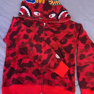 Bape hoodie - Säljer en cool röd camo hoodie från BAPE med hajmotiv på huvan. Hoodien har dragkedja och broderade bokstäver i olika färger. Perfekt för dig som vill sticka ut med en unik stil!