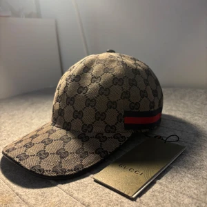 Beige gucci keps - Snygg keps från Gucci med klassiskt mönster i beige och brunt. Kepsen har en röd och grön randdetalj på sidan. Perfekt för att ge din outfit en lyxig touch. Storlek ”S”