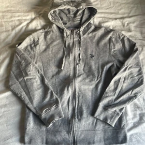 Grå hoodie från U.S. Polo Assn. - Säljer en stilren grå hoodie från U.S. Polo Assn. med dragkedja och huva. Tröjan har ett diskret tryck av logotypen på bröstet och är tillverkad i mjukt material för extra komfort. Tröjan är i storlek S och har inga skador eller andra skavanker. 