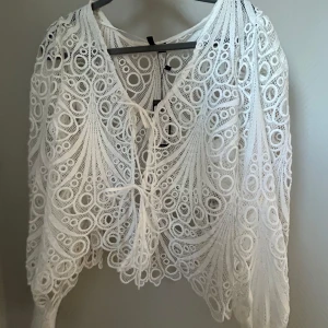 Vit spetsblus från Vero Moda - Säljer en elegant vit spetsblus från Vero Moda med v-ringning och knytning framtill. Blusen har ett unikt cirkulärt mönster och långa ärmar, perfekt för en stilfull look. Passar bra till både jeans och kjol. 🌟