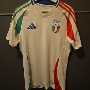 Italien EM 2024 fotbollströja  - Säljer en vit fotbollströja från Adidas med Italiens flagga och tre stjärnor på bröstet. Den är köot på unisport i Stockholm och är äkta. Tröjan har korta ärmar med röd och grön detaljering på sidorna och axlarna. Perfekt för fotbollsfans! 🇮🇹