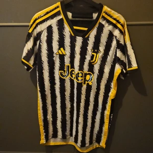 Juventus fotbollströja med Vlahović 9 - Säljer en Juventus fotbollströja från Adidas med Vlahovićs namn och nummer 9 på ryggen. Tröjan har ett unikt svartvitt randigt mönster med gula detaljer och korta ärmar. Perfekt för fans av laget!