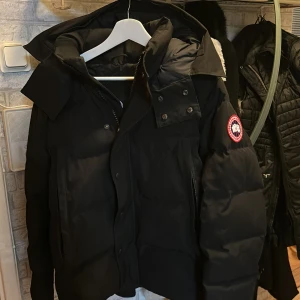 Svart dunjacka från Canada Goose - Säljer en svart dunjacka från Canada Goose med deras ikoniska märke på ärmen. Jackan har en stor huva och stängs med dragkedja och knappar. Perfekt för kalla vinterdagar.