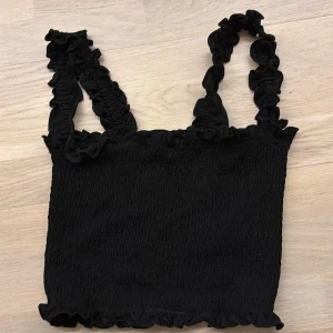 Svart ribbad linnetopp - Säljer en svart ribbad linnetopp med volangdetaljer på axelbanden och längs kanterna. Perfekt för en sommar look. Linnet är cropped men ser perfekt ut till en kofta! 