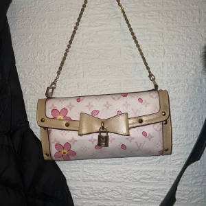 Blommig axelväska med rosett - Säljer en söt axelväska med blommigt mönster i rosa och beige. Väskan har en dekorativ rosett och ett lås framtill. Den har en kedjerem i guld som ger en elegant touch.       Kedjan har jag själv satt på.