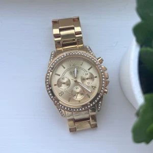 Guldfärgad klocka med strassdetaljer - Snygg guldfärgad klocka från O.N.E med strass runt urtavlan och länkar. Urtavlan har flera små visare och en elegant design. Perfekt för att ge en touch av glamour till din outfit. Sparsamt använd. Extra länkar för att göra armbandet längre följer med. Önskar du veta exakta mått? Tveka inte att höra av dig!