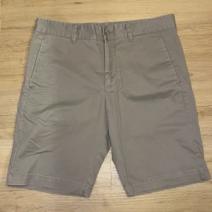 Beige shorts från Peak Performance - Snygga beige shorts från Peak Performance med knapp och dragkedja framtill. De har två bakfickor med knappar och en stilren design. Perfekta för en avslappnad stil.