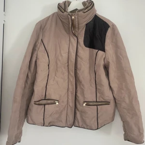 Beige quiltad jacka med svarta detaljer - Snygg beige jacka med svarta detaljer och dragkedjor. Jackan har en hög krage och två framfickor med dragkedja. Perfekt för kyligare dagar.