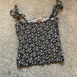 Blommig svart linnetopp från Stories - Säljer en söt svart linnetopp från Stories med vita blommor. Toppen har smala axelband som knyts. Ser liten ut men är väldigt stretchig 