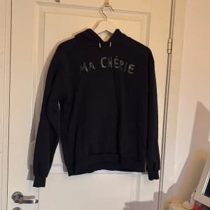 Svart hoodie från Gina Tricot - Säljer en svart hoodie från Gina Tricot med texten 'MA CHÉRIE' i glittriga stenar på framsidan. Tröjan har en klassisk huva med dragsko och långa ärmar. Perfekt för en avslappnad stil.