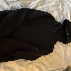 Svart hoodie från C.P. Company Storlek S - Säljer en stilren svart hoodie från C.P. Company. Den har en klassisk design med dragsko i huvan och en liten logga på ärmen. Perfekt för en avslappnad look.