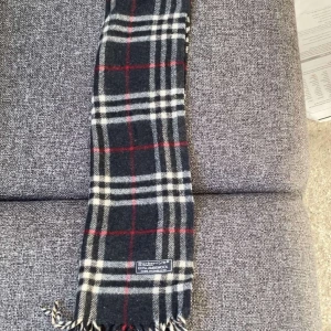 Rutig halsduk från Burberry - Snygg rutig halsduk från Burberry i klassiska färger. Den har en bas i mörkgrått med vita och röda rutor. 