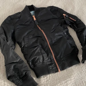 Svart bomberjacka från Alpha Industries - Säljer en snygg svart bomberjacka från Alpha Industries. Jackan har en rosefärgad dragkedja och en ficka med dragkedja på ärmen. Den är tillverkad i syntetmaterial och har ribbade muddar vid ärmslut och nederkant. Perfekt för en cool och avslappnad stil.