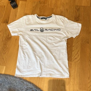 Vit t-shirt från Sail Racing - Säljer en vit t-shirt från Sail Racing med svart text på bröstet. T-shirten har en klassisk rund halsringning och korta ärmar. Perfekt för en avslappnad stil.