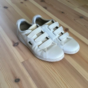 Vita sneakers från Lacoste - Snygga vita sneakers från Lacoste med tre praktiska kardborreband för enkel på- och avtagning. Skorna har en klassisk design med den ikoniska krokodilloggan på sidan. Perfekta för en stilren look.