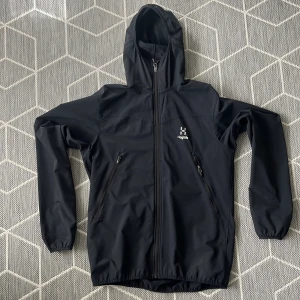 Svart vindjacka från Haglöfs - Säljer en svart vindjacka från Haglöfs med huva och dragkedja framtill. Jackan har två praktiska fickor med dragkedjor och är perfekt för utomhusaktiviteter. Den är långärmad och har en stilren design.