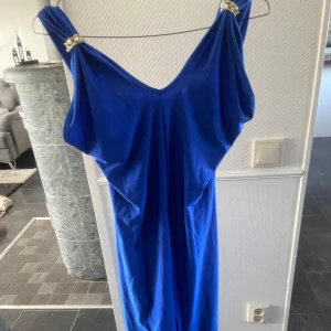Blå långklänning med gulddetaljer - Snygg blå långklänning med v-ringad design och gulddetaljer vid axelbanden. Perfekt för en elegant kvällslook. Klänningen har en smickrande passform och är tillverkad i ett mjukt material som ger en fin drapering.