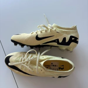 Gul/beige och svarta fotbollsskor från Nike - Snygga fotbollsskor från Nike i beige med svarta detaljer och snörning. Skorna har en modern design med tydlig Nike-logga och är perfekta för spel på gräs. De har en lätt och smidig passform som ger bra grepp och kontroll på planen.