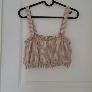 Beige och vit randig linnetopp från Gina Tricot - Säljer en söt beige och vit randig linnetopp från Gina Tricot. Toppen har breda axelband och en elastisk kant upptill och nedtill för en bekväm passform. Perfekt för varma sommardagar!