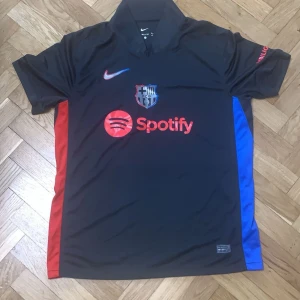 Svart fotbollströja med röda och blå detaljer - Säljer en svart fotbollströja från Nike med röda och blå detaljer på sidorna. Tröjan har korta ärmar och ett tryck av Spotify-loggan på framsidan. Perfekt för fotbollsfans som vill ha en stilren look.