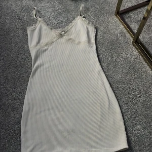 Vit ribbad klänning från H&M Divided - Säljer en stilren vit ribbad klänning från H&M Divided. Klänningen är ärmlös med smala axelband och har en söt spetsdetalj vid halsringningen. Perfekt för en avslappnad look eller att klä upp med accessoarer.