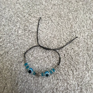 Blått armband med ögonmotiv - Snyggt armband med blå pärlor och ögonmotiv. Armbandet har ett justerbart svart snöre för perfekt passform. Perfekt för att ge en unik touch till din stil.