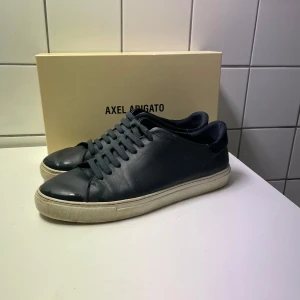Mörkblåa sneakers från Axel Arigato - Snygga mörkblåa sneakers från Axel Arigato med en stilren design. Skorna har en vit sula och är tillverkade i skinn med snörning framtill. Perfekta för en casual look. Skorna har visaa märken på sulan vilket drar ner priset såpass mycket. Box och påse medföljer.