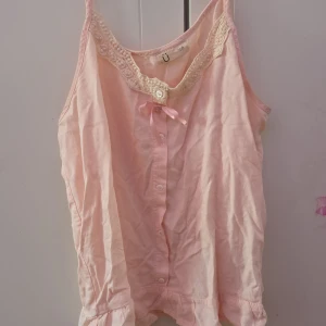 Rosa linne med spetsdetaljer - Söt rosa linne med spetsdetaljer vid halsringningen och en liten rosett framtill. Linnet har knappar längs framsidan och är perfekt för varma dagar.