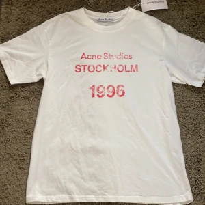 Vit t-shirt från Acne Studios - Säljer en vit t-shirt från Acne Studios med tryck i rött som säger 'STOCKHOLM 1996'. Perfekt för en stilren och avslappnad look. T-shirten har en klassisk passform och rund halsringning. Ganska stor för S.