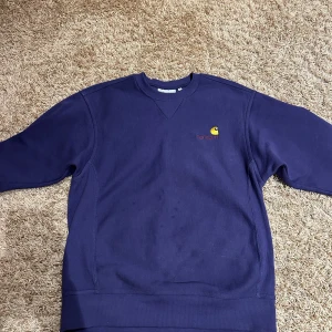 Mörkblå sweatshirt från Carhartt - Säljer en snygg mörkblå sweatshirt från Carhartt med broderad logga i gult och rött på bröstet. Tröjan har en klassisk rund hals och långa ärmar. Perfekt för en avslappnad stil.