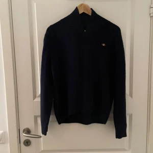 Mörkblå stickad tröja från GANT - Säljer en stilren mörkblå stickad tröja från GANT. Tröjan har en hög krage och ett diskret märke på bröstet. Perfekt för kyligare dagar med sin ribbade struktur och långa ärmar. Passar både till vardags och mer uppklädda tillfällen.