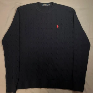 Mörkblå stickad tröja från Ralph Lauren - Säljer en stilren mörkblå stickad tröja från Ralph Lauren med kabelstickat mönster. Tröjan har en rund halsringning och ett rött broderat märke på bröstet. Tröjan passar runt 180 eller 175